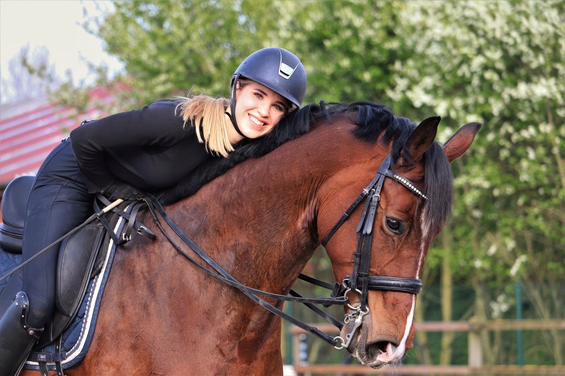 Lena Lüttmer-Strathmann mit Reithelm sitzt auf einem braunen Pferd, lehnt sich lächelnd auf seinen Hals und schaut in die Kamera. Im Hintergrund blühende Bäume.